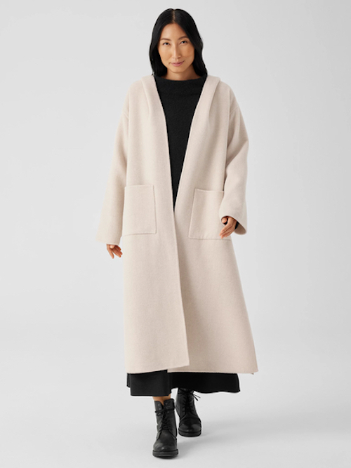 A woman with black hair wears an elegant, cream-colored coat going nearly to her ankles over a long black dress and black ankle-height boots.