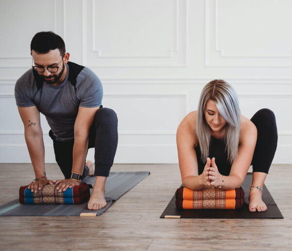 A bearded, dark-haired man and a silver-haired woman, both in their 20s or 30s, practice yoga on mats in a room with white walls and a wooden floor. Each uses a colorful, patterned Zafuko cushion to support their yoga poses.